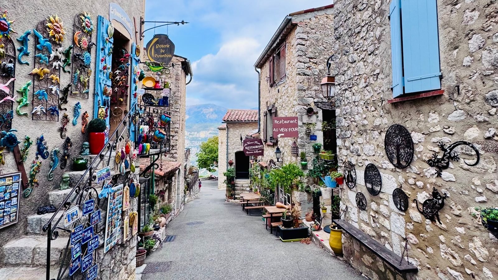 A village street in Gourdon, shop walls hung with ceramics, blue shutters, the mountains at the end of the lane.